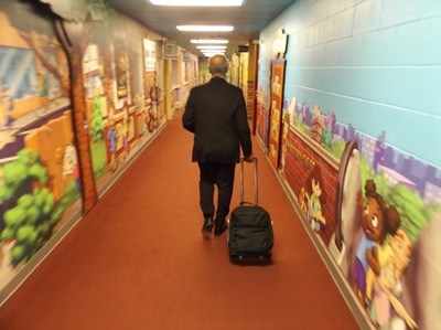 Man walking down hall pulling luggage on wheels.
